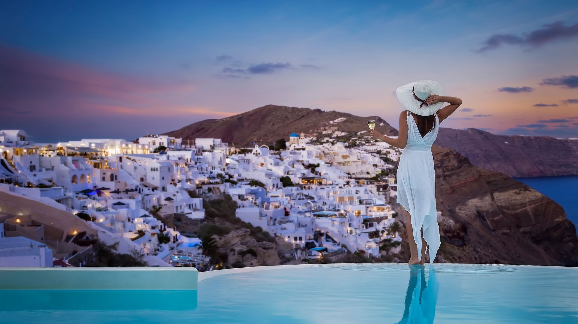 A woman in a white dress stands by the swimming pool holding a wine glass and enjoys the evening view of Oia during her 9 days in Greece.