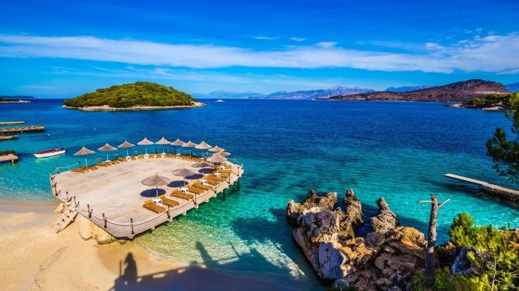 A coastal scene with a wooden deck, sun loungers and umbrellas extending over clear, turquoise waters and islands in the distance.