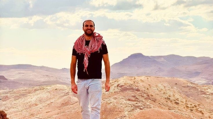 A bearded man with a shemagh around his neck stands against arid valley