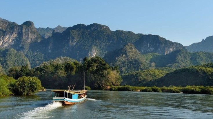 A tourist boat sails on the Mekong River with verdant mountains in the background, in Luang Prabang, Laos. Embark on a tourist cruise for a tour of the Mekong River in Luang Prabang.