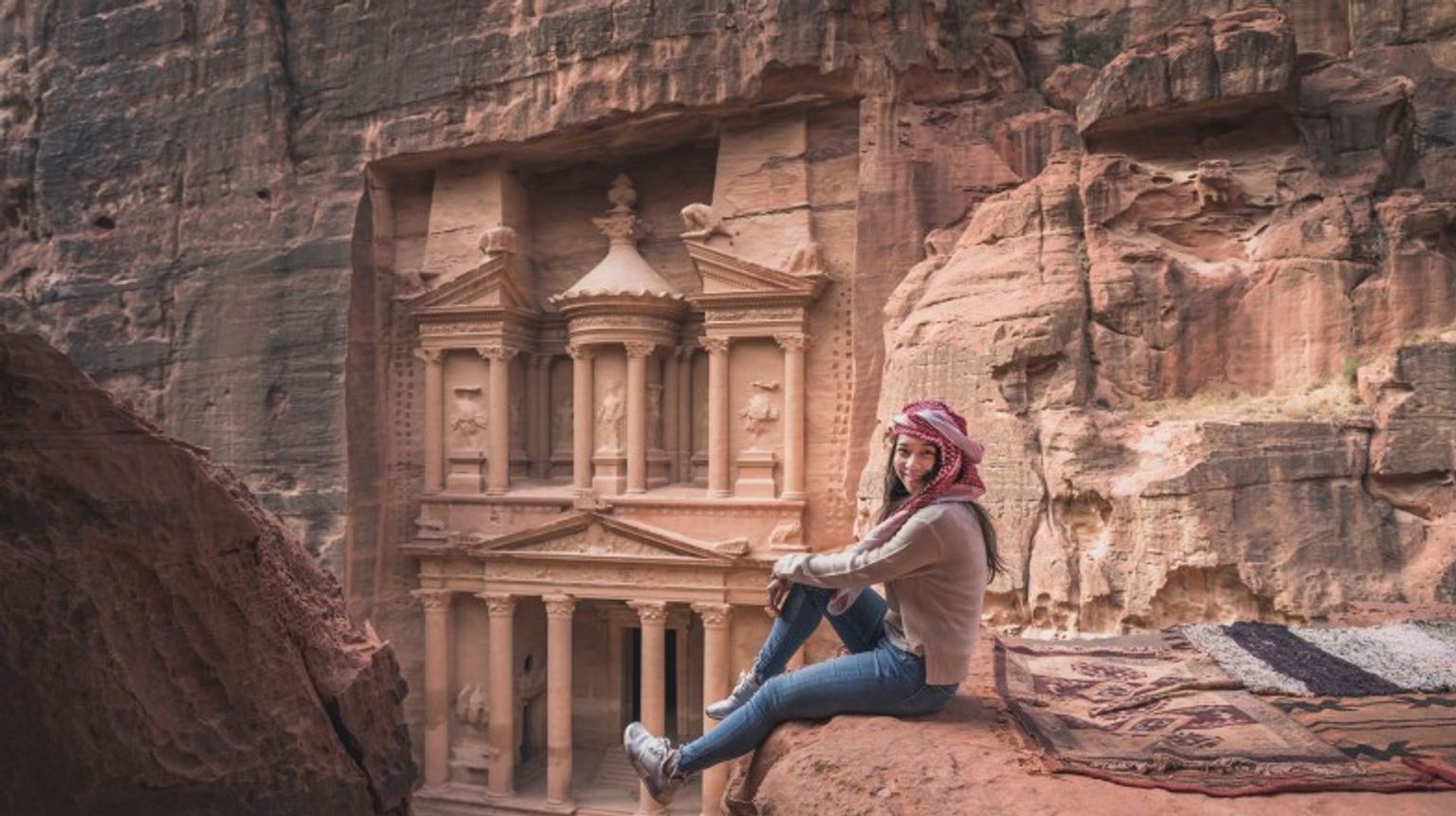 Visit Petra and explore the UNESCO World Heritage site during your 4 days in Jordan, where ancient ruins and stunning views await. A tourist girl smiling sitting on the rock in front of Treasury during her 4 days in Jordan.