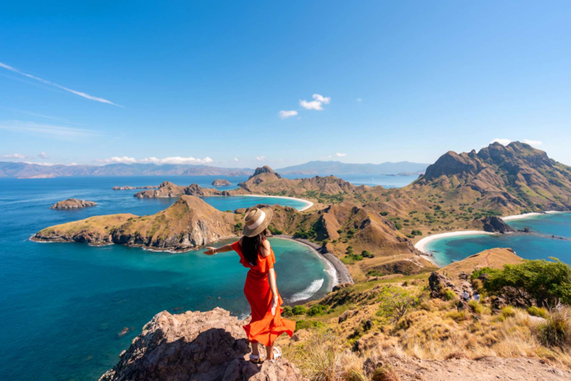 A woman in orange dress stand on top of a small hill surrounded by sea and lines of other small hills in Indonesia