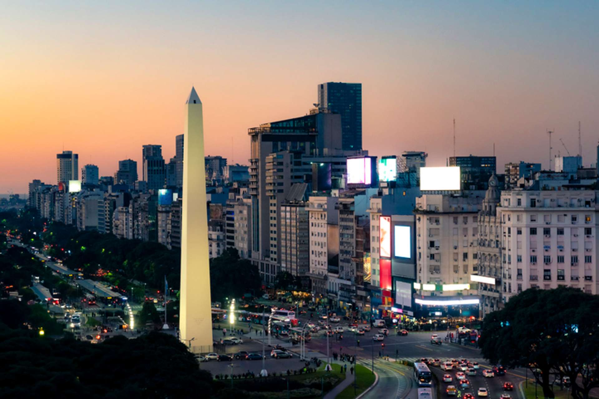 A beige, obelisk structure stands at the conjunction of a busy street with buildings lined on one end