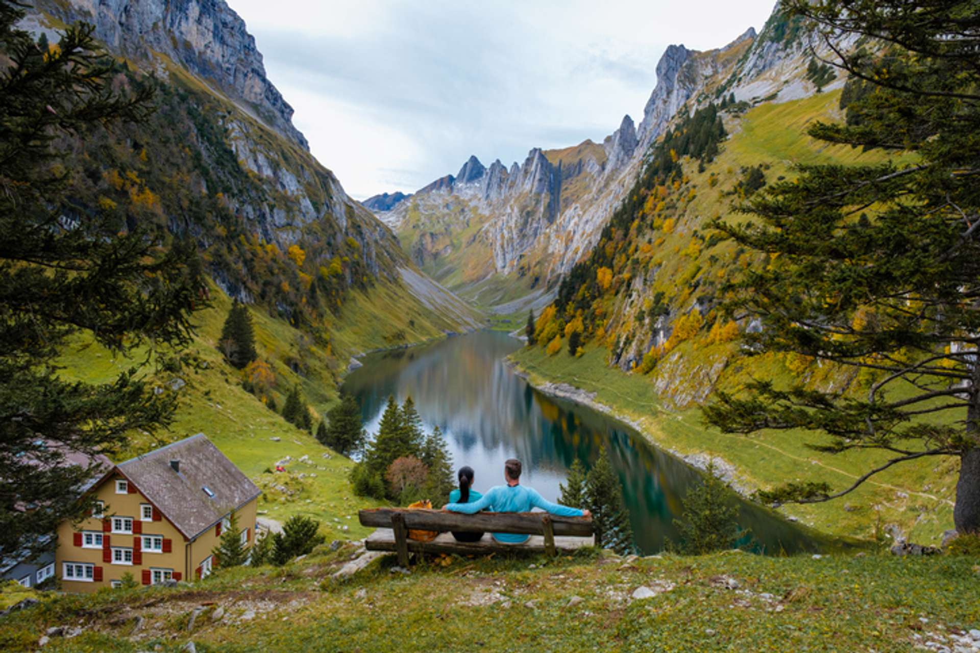 A couple sits on a bench overlooking a view of a lake surrounded by rocky mountains