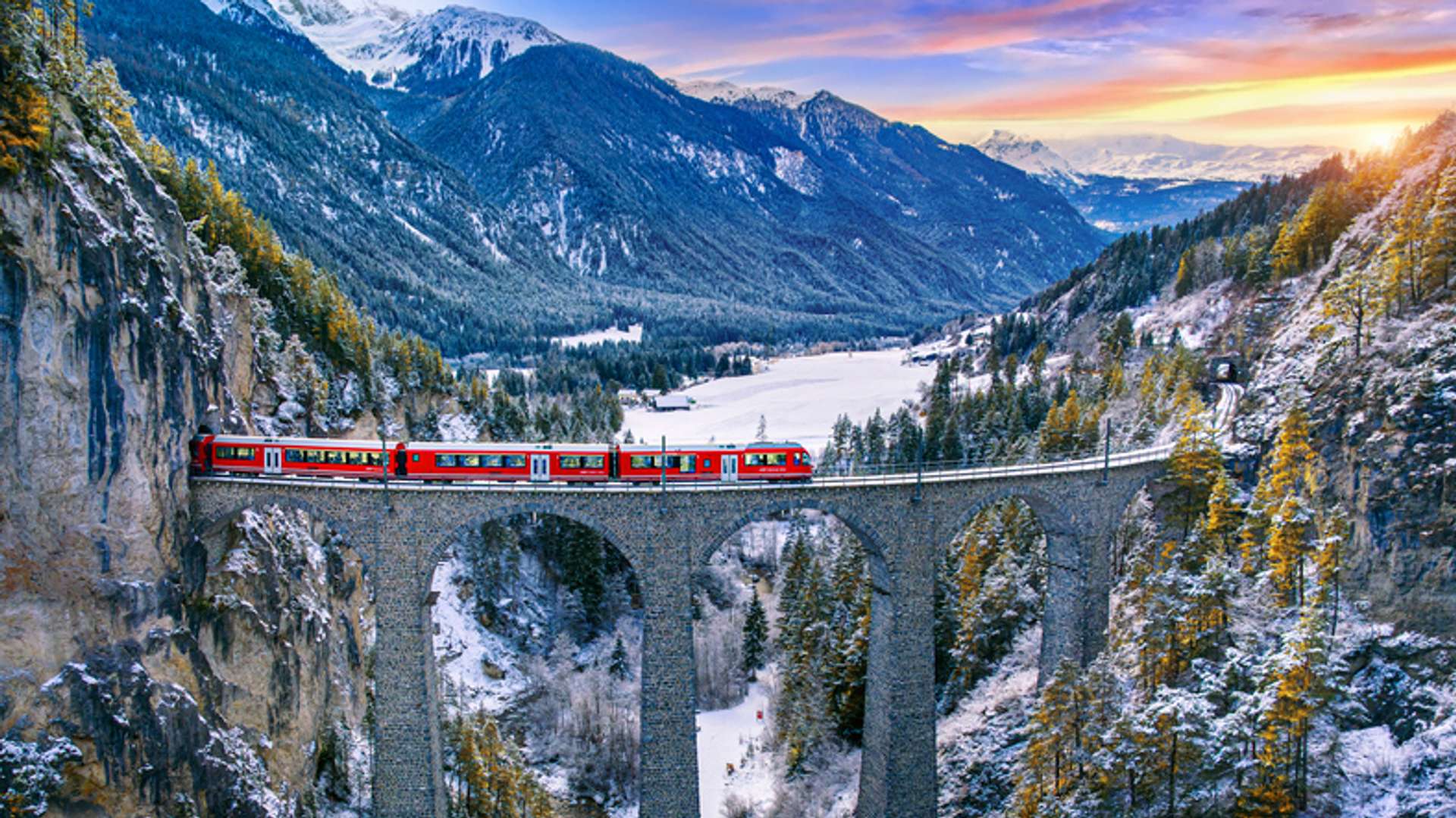 Red train going through a train track on a bridge with snowy mountains and rivers in view