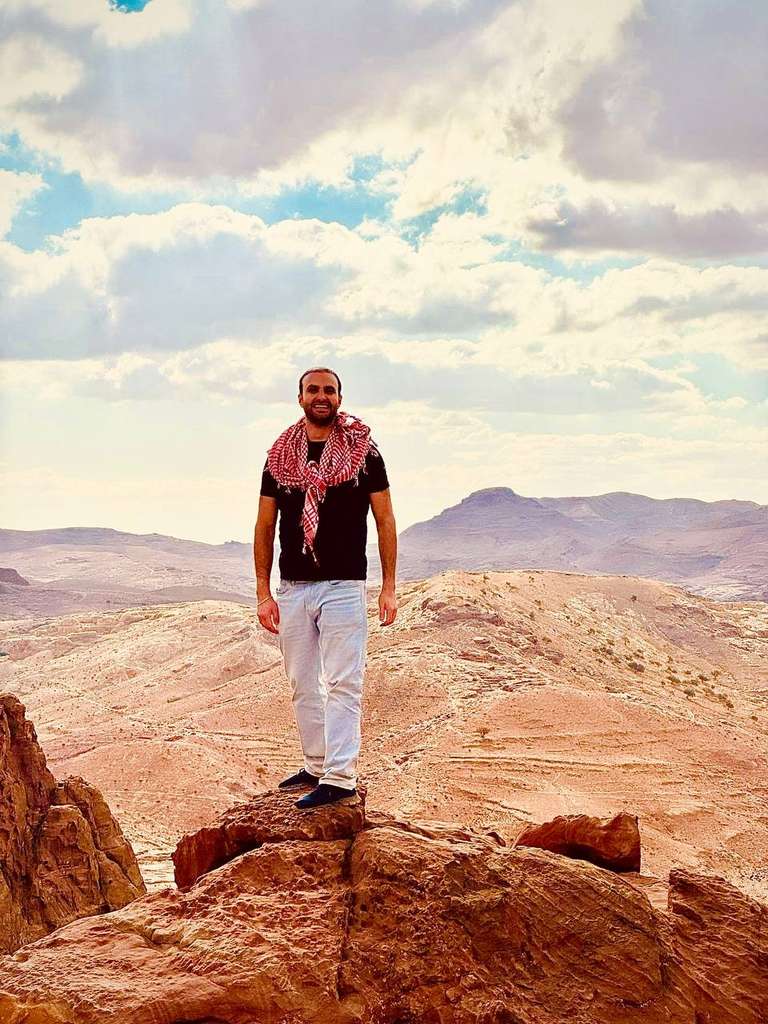 A young man with a scarf around his shoulders stands in a cliff around Petra Monastery