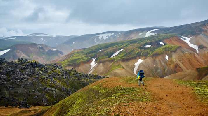 A hiker walking through colorful mountains of Landmannalaugar against a dark sky in Iceland.