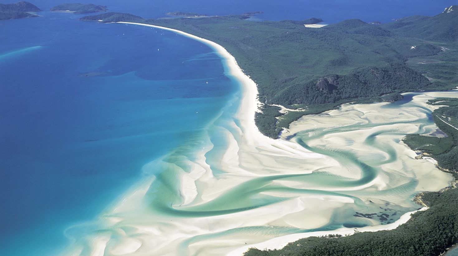 Aerial view of Whitehaven Beach with white waves and blue water surrounding hills. 