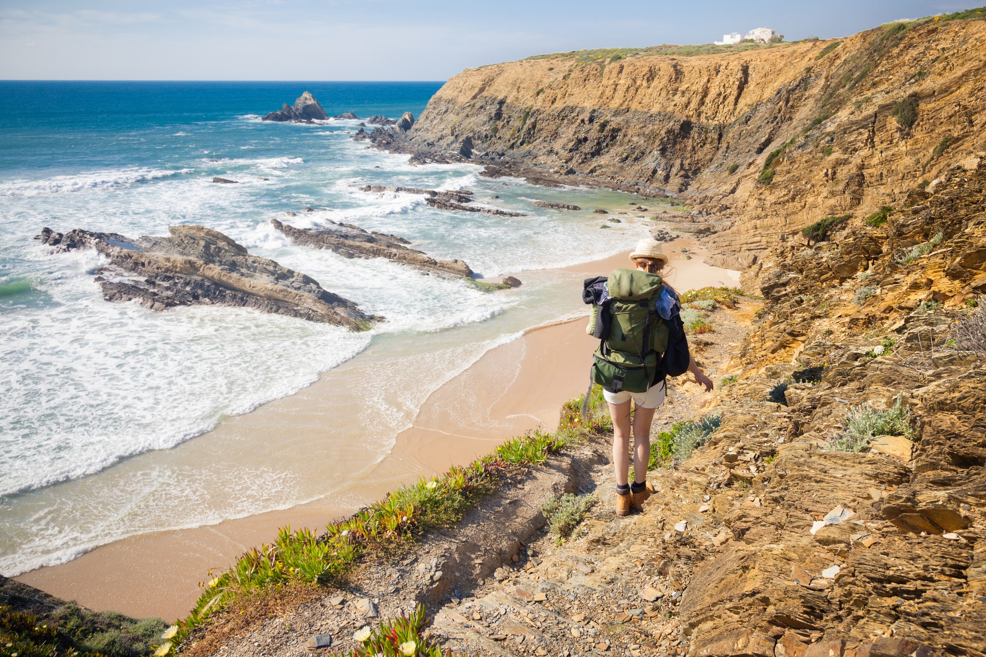 The famous Rota Vicentina is made up of two hiking trails: the Fisherman’s Trail and the Historical Way. A woman hiking down one of the many rugged, coastal trails in Portugal with the ocean & beach in view.