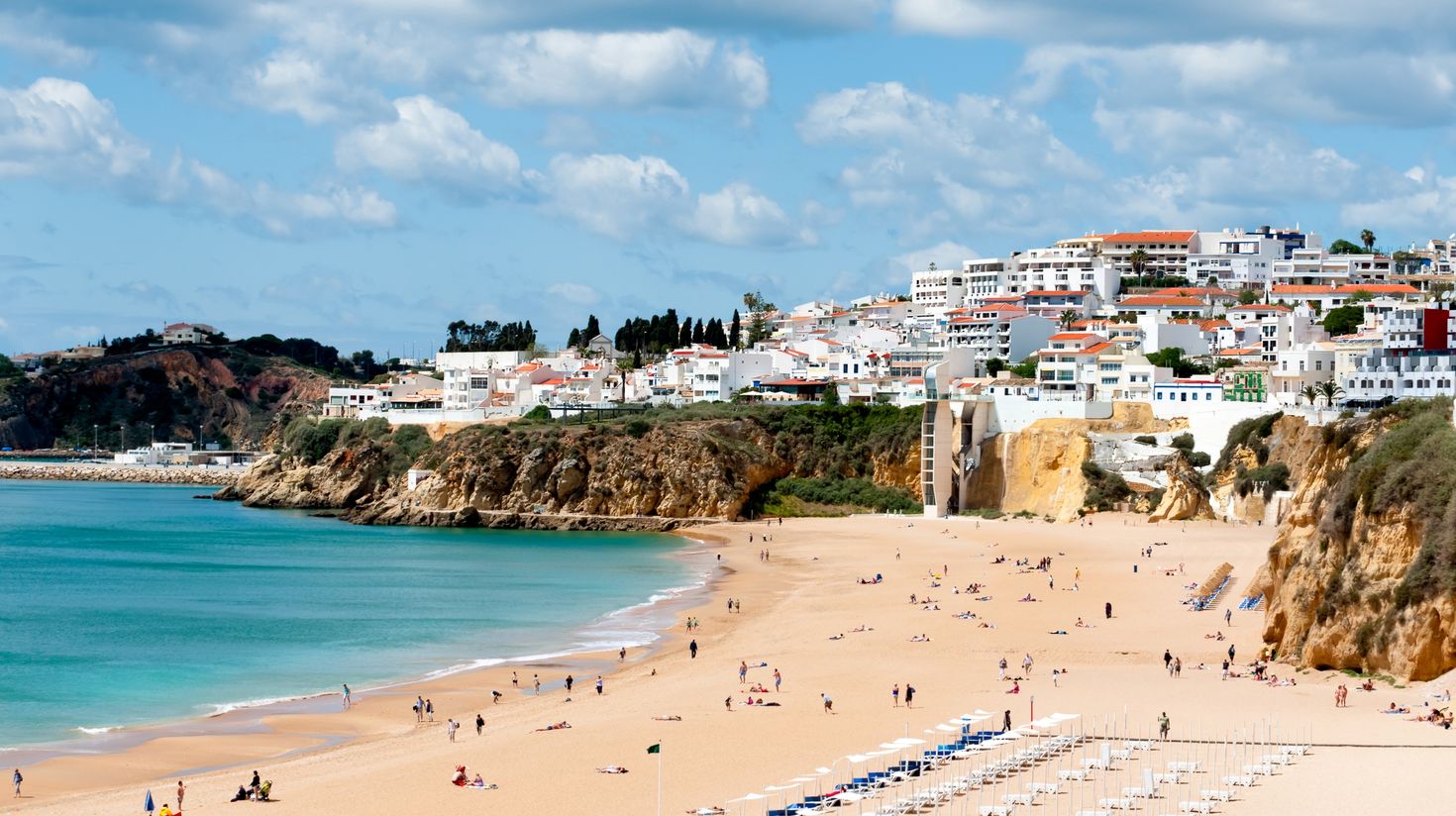 A rocky coastline town with white-washed houses and a strip of sandy beach with people.