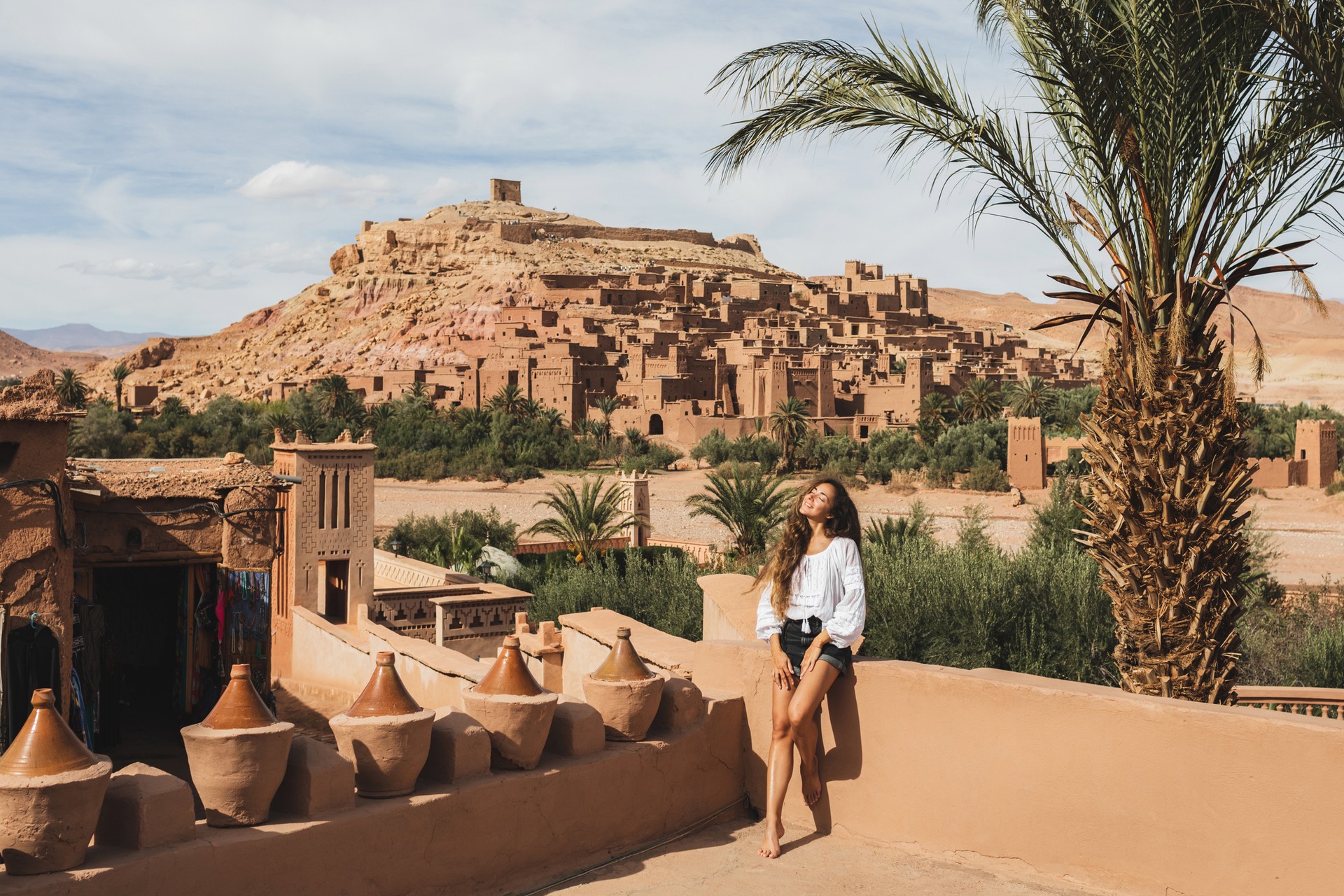 Discover the ancient beauty of Ait-Ben-Haddou during your 8 days in Morocco, where this UNESCO World Heritage site enchants with its stunning kasbahs and desert land A happy tourist girl posing in front of Ait-Ben-Haddou during her 8 days in Morocco.