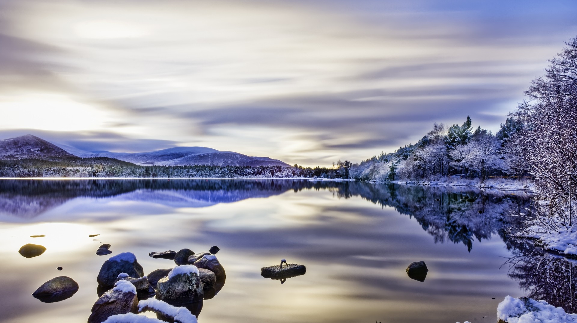 Discover Scotland in January, with a day full of breathtaking scenery. Soft clouds sweep across the sky, snow on trees, rocks and water gives way to reflections Beautiful winters day with soft clouds, snow on trees and rocks, reflections on calm water in Scotland in January.