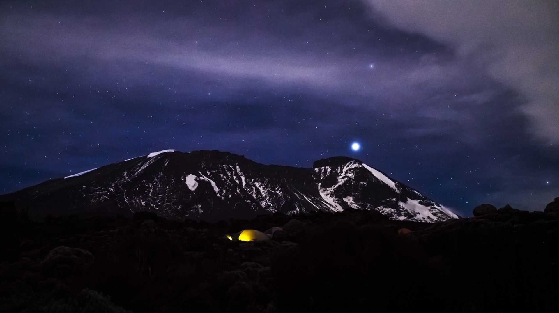 An evening sight of a snow covered mountain with a glowing yellow tent at its base.