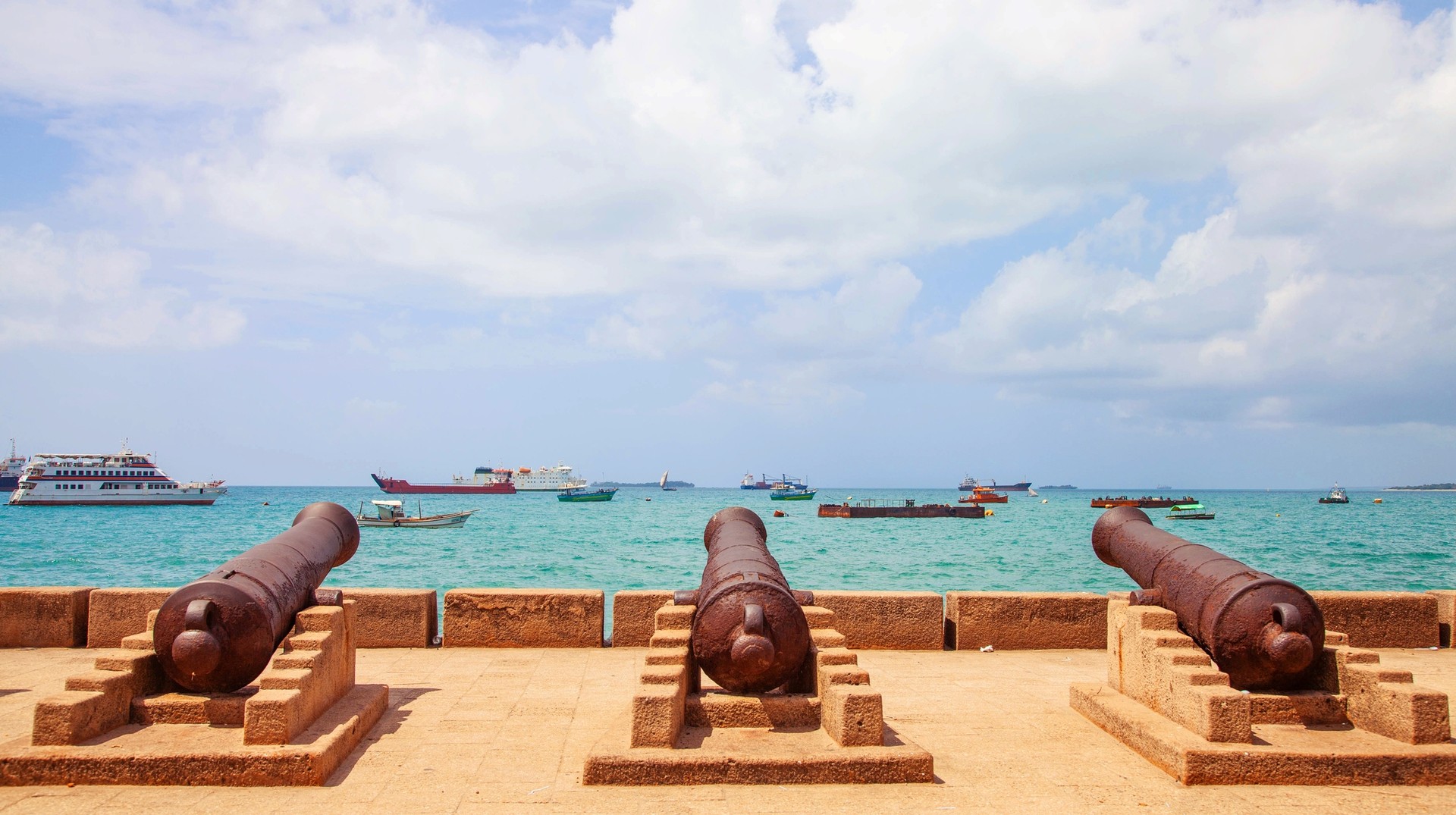 One of the best things to do in Zanzibar is to hop on a boat and get to all significant destinations, including the historically significant Stone Town. Three antique cannons lined up next to each other and positioned towards turquoise sea with ships and boats—one of the best things to do in Zanzibar.