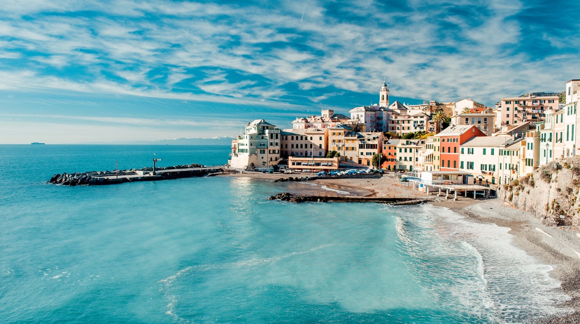 A coastal town with white, red buildings next to a blue sea with waves washing into the beach.