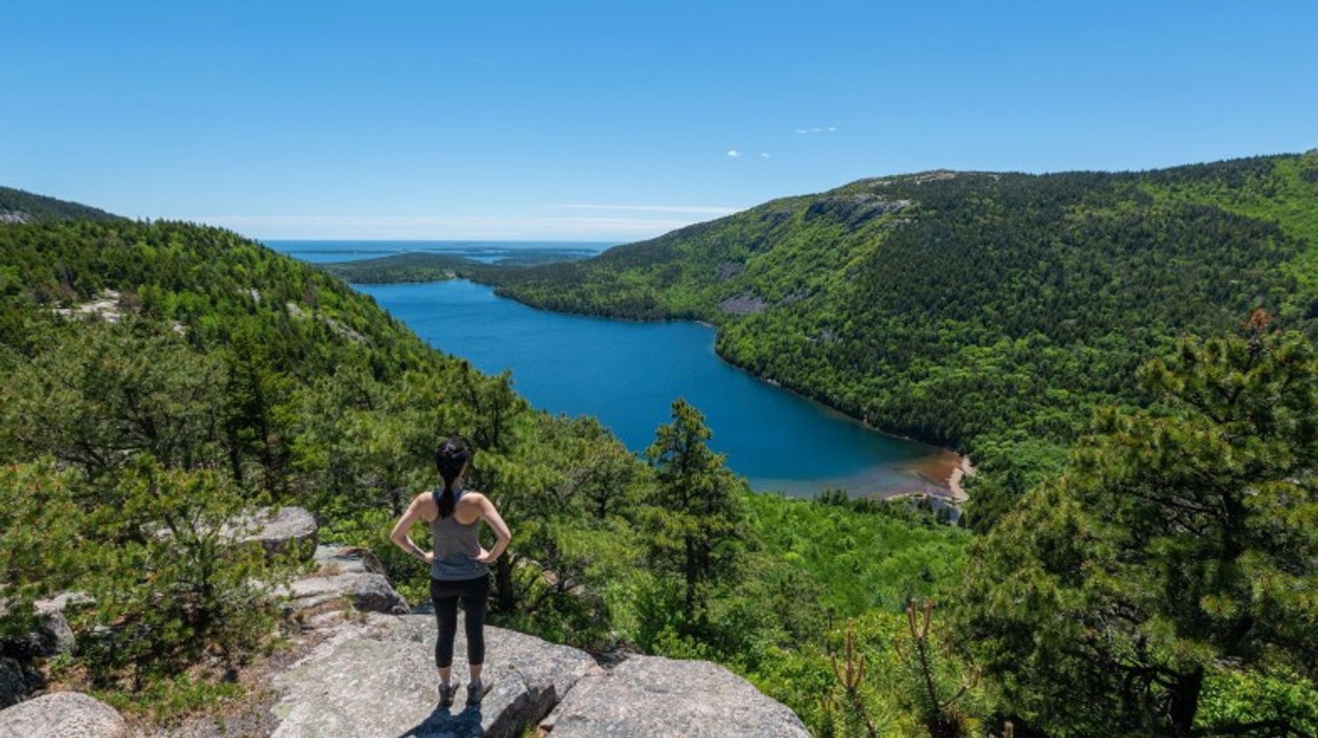 Immerse yourself in the breathtaking beauty of Acadia National Park, where rugged coastlines, serene lakes, and lush forests during your 12 days in The USA. A woman enjoying the view of Acadia National Park on a clear day during her 12 days in The USA.