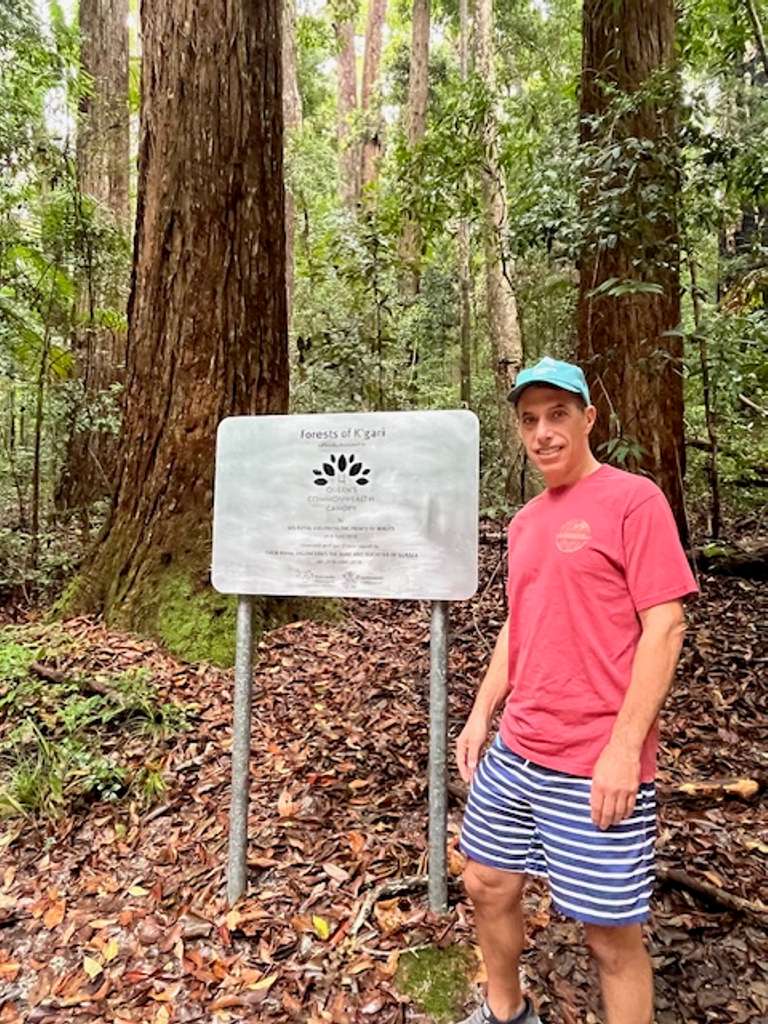 A man in red tshirt and shorts stands next to a notice board in a forest
