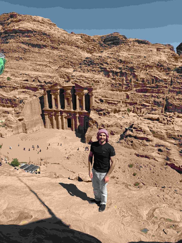 A man wearing a scarf stands on top of a cliff with a view of an ancient building carved into a mountain
