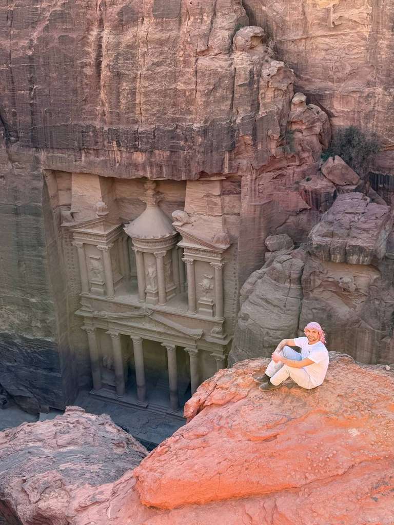 A man with a scarf on his head sits on a cliff with the view of an ancient building carved into a mountain