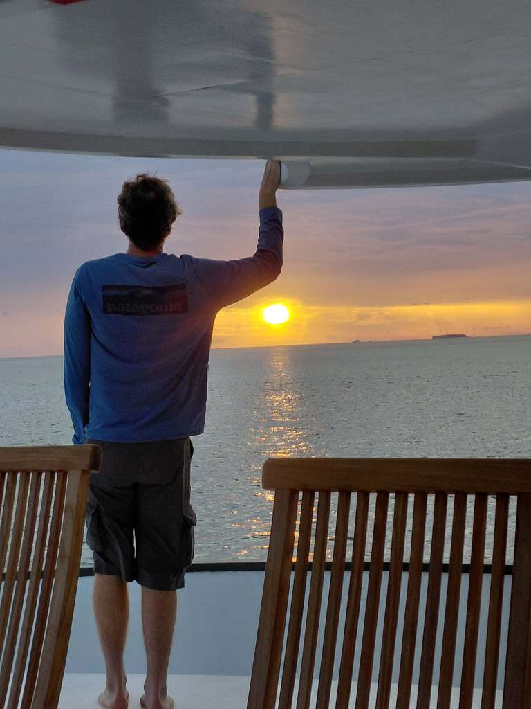 A man stands on a yacht with his back to the camera looking at a sunset over the sea